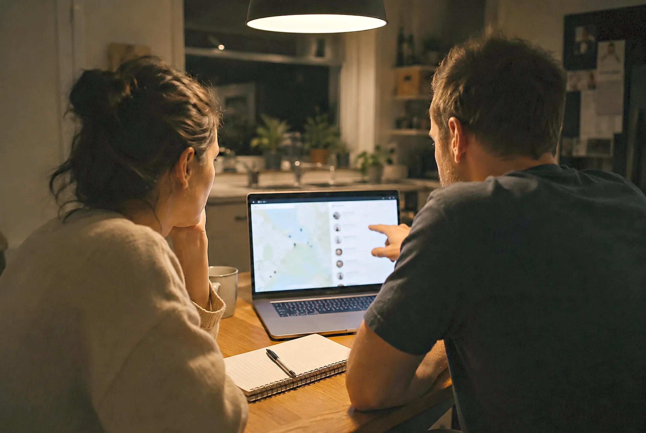 A couple at a kitchen bench in warm evening light look through local trade businesses on a laptop together.
