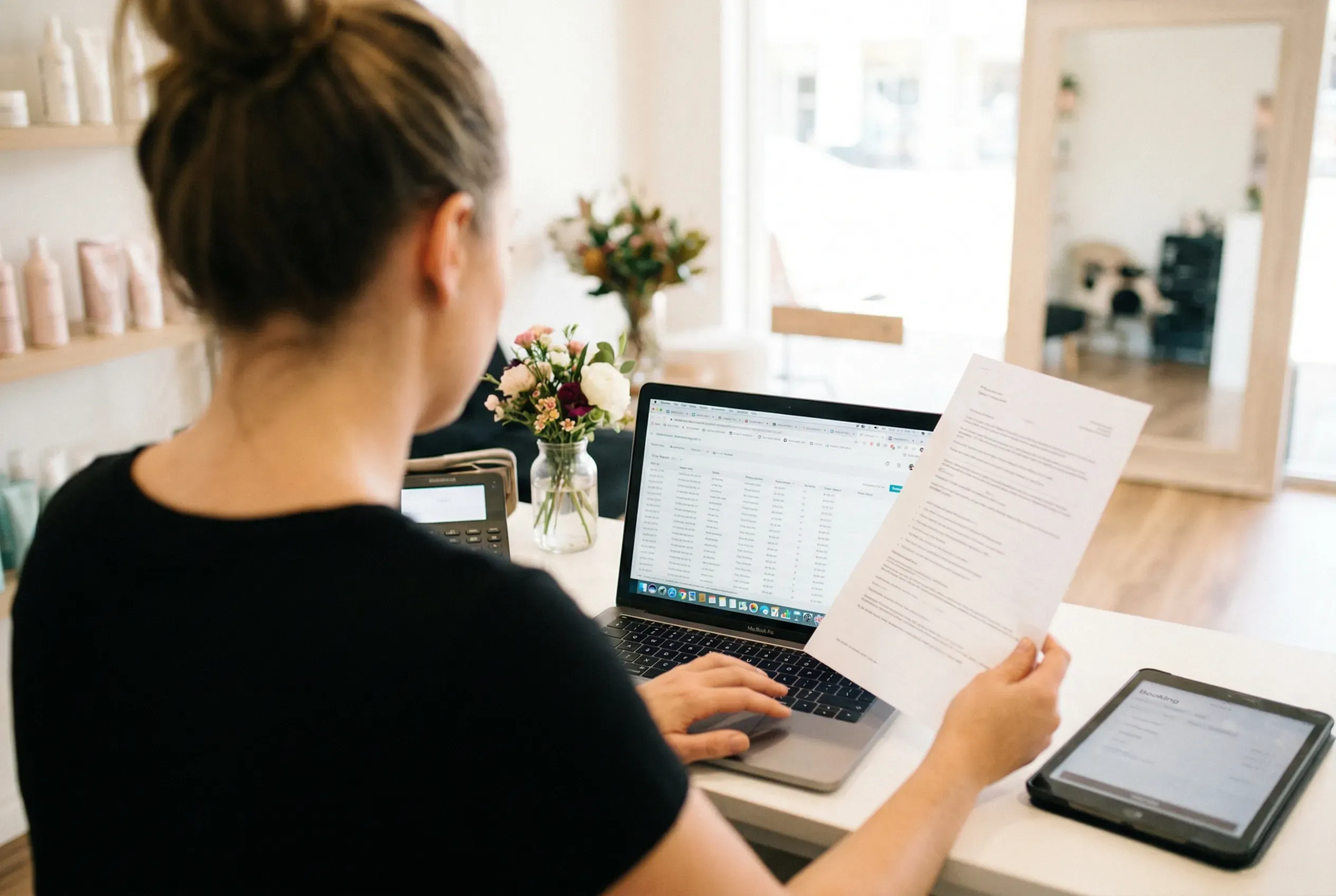 Salon owner comparing a printed report against a spreadsheet on her laptop at the front counter