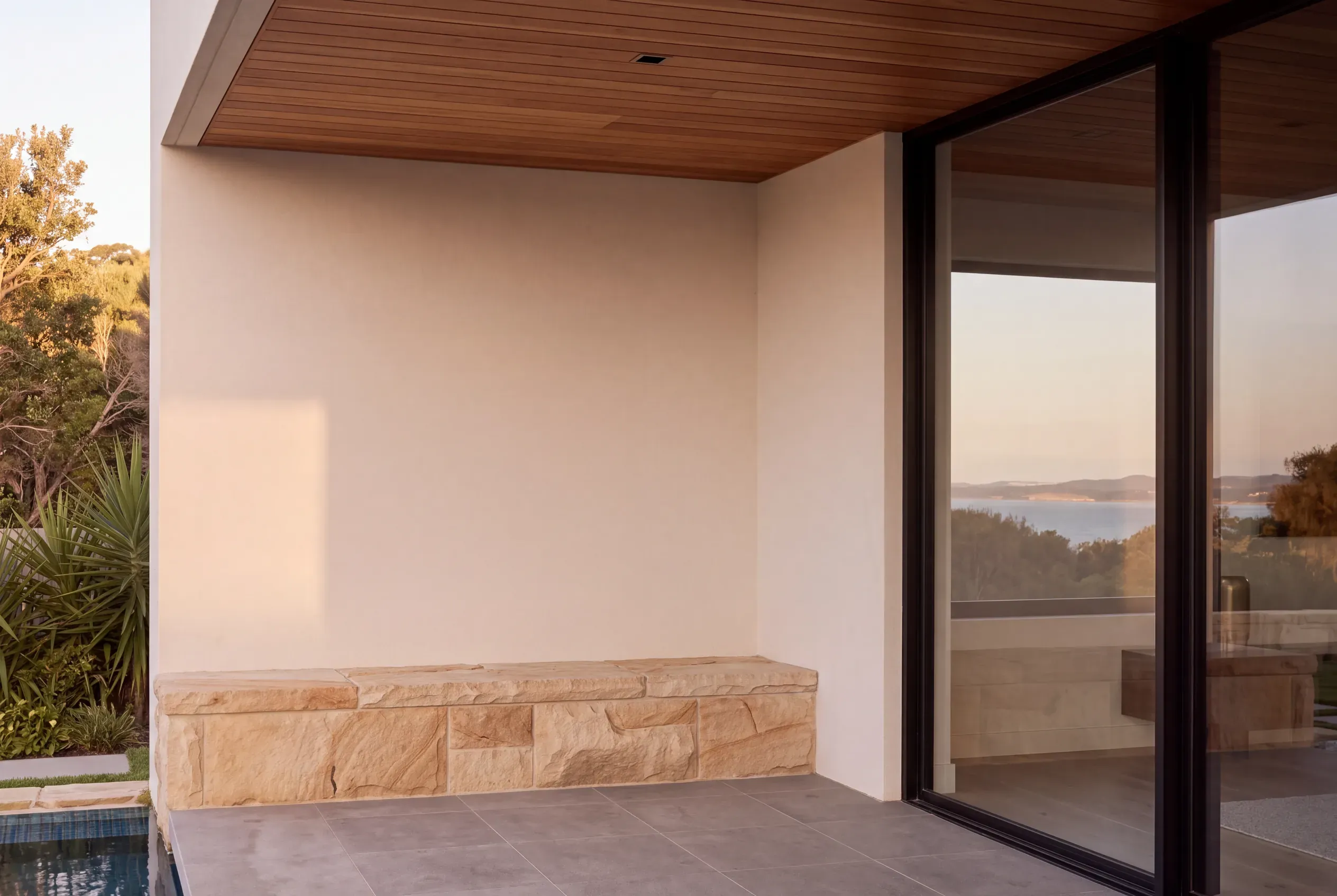 A close view of a sandstone bench base meeting smooth warm white render under a timber-lined soffit, beside a black-framed glass sliding door reflecting the water.